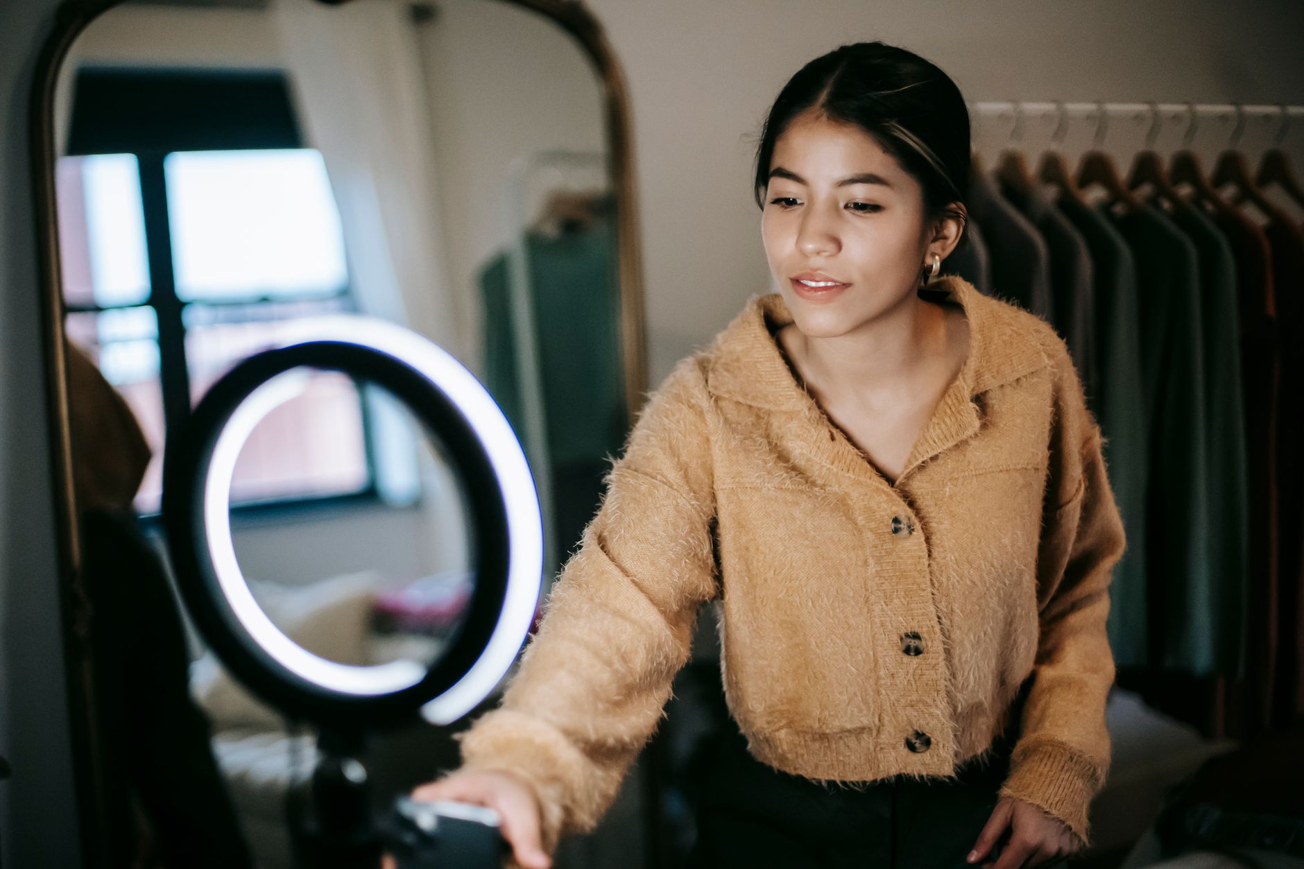 Joven sonriendo frente a un aro de luz con el móvil, en un cuarto con espejo y una barra de ropa